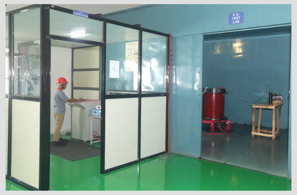 A partial-discharge test bay signed P.D. TEST LAB holding a red high-voltage test set on a wheeled frame, with an operator in a hard hat working at instruments in the separate glazed P.D. control room alongside it.